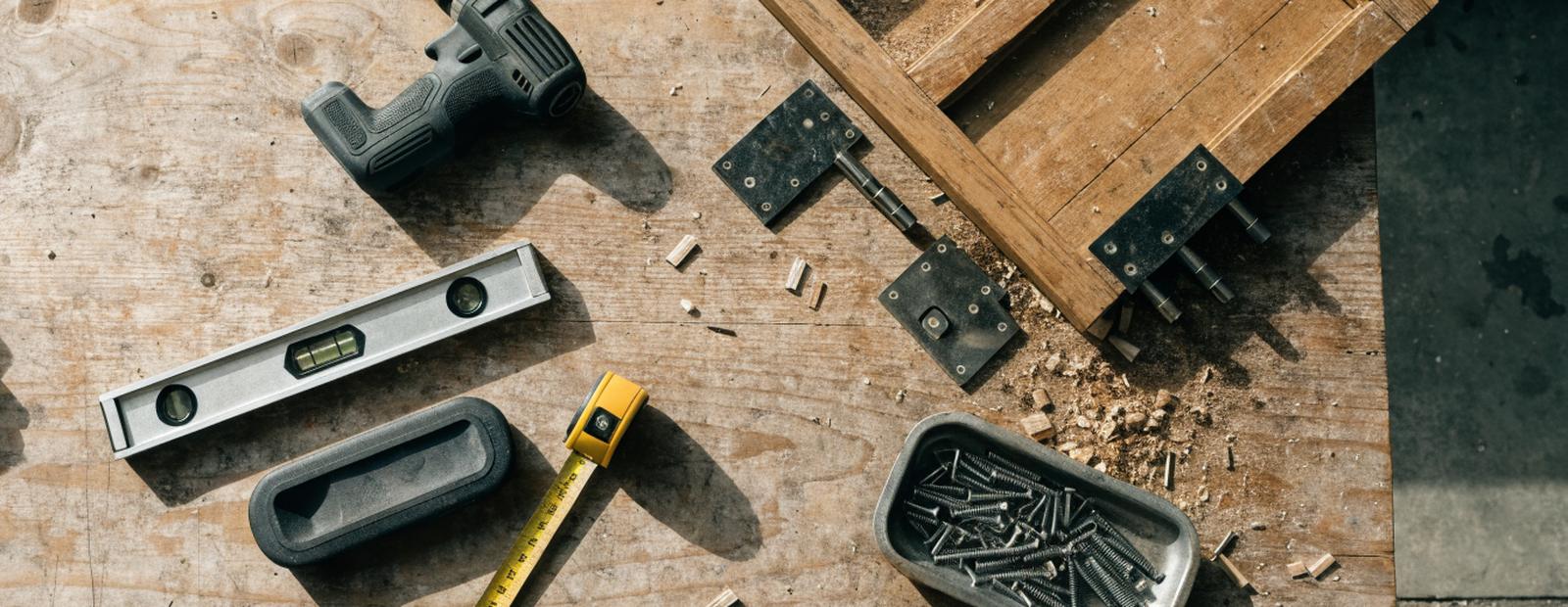 A workbench flat-lay of a cordless drill, level, tape measure and screws beside a partially disassembled cabinet door