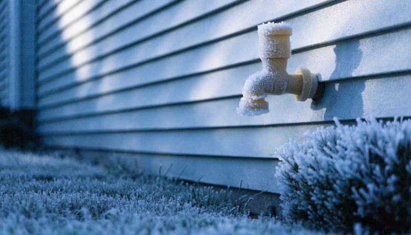 An outdoor spigot on house siding wearing an insulated foam freeze cover, heavy frost on the lawn below